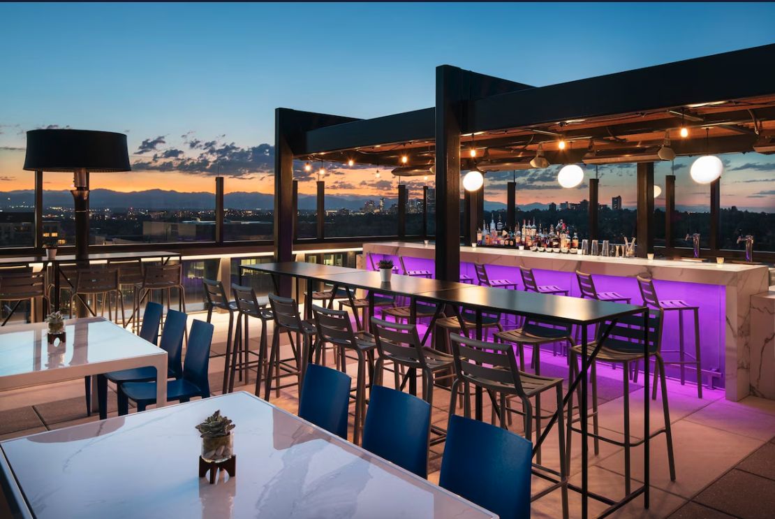 Rooftop bar at dusk with modern seating, purple-lit bar, city skyline, and mountain views in Cherry Creek, Denver.